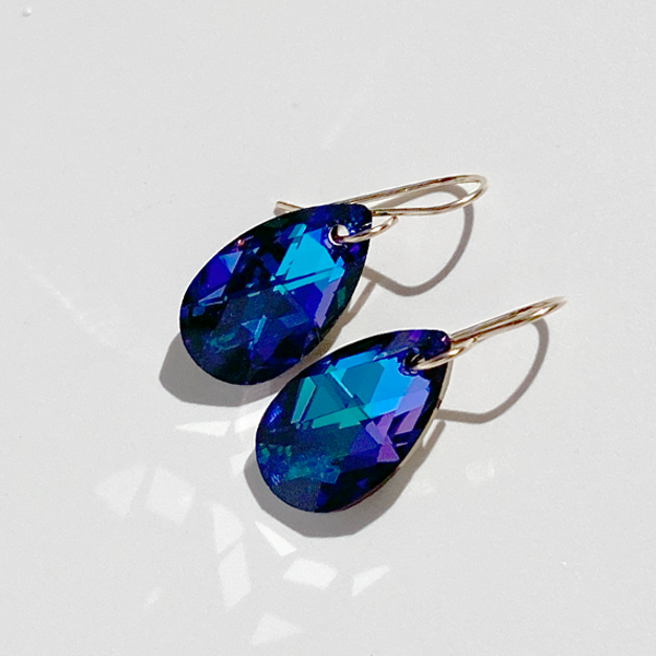 A pair of elegant pear-shaped crystal earrings with blue and purple hues, attached to gold-colored hardware, displayed against a white background.
