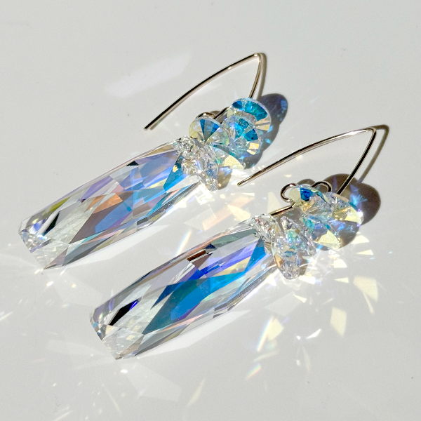 A pair of long crystal earrings with iridescent stones and gold filled hardware, laid out on a white surface.