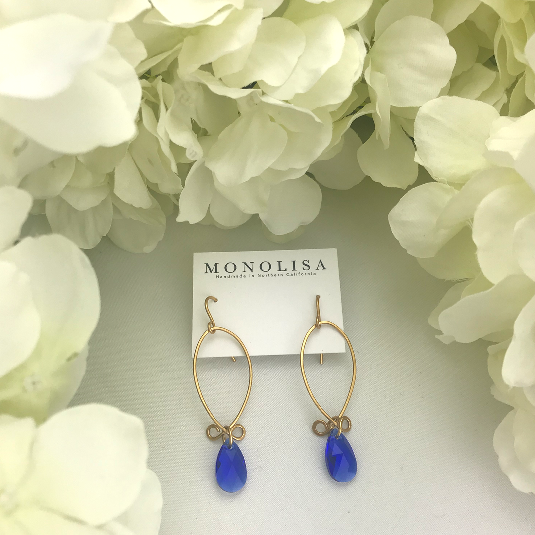 A pair of gold-colored hoop earrings with blue dangling stones, displayed on a card with the brand name 'MONOLISA' written on it, set against a background of white hydrangea flowers.